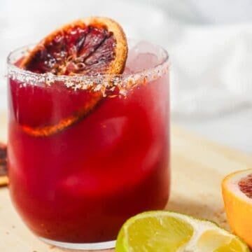 A glass of red cocktail with ice, garnished with a blood orange slice, next to lime and orange halves.