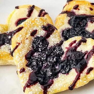 Heart-shaped pastries with dark berry jam, perfect for lovers of blueberry recipes, dusted with powdered sugar and arranged on a white plate.