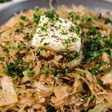 Close-up of sautéed cabbage topped with a pat of butter, fresh herbs, and seasonings on a plate.