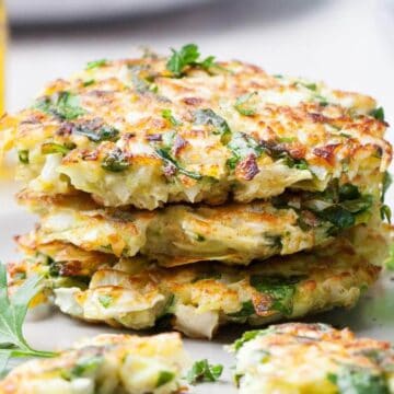 Three zucchini fritters stacked on a plate with parsley garnish and a small bowl of dipping sauce nearby.