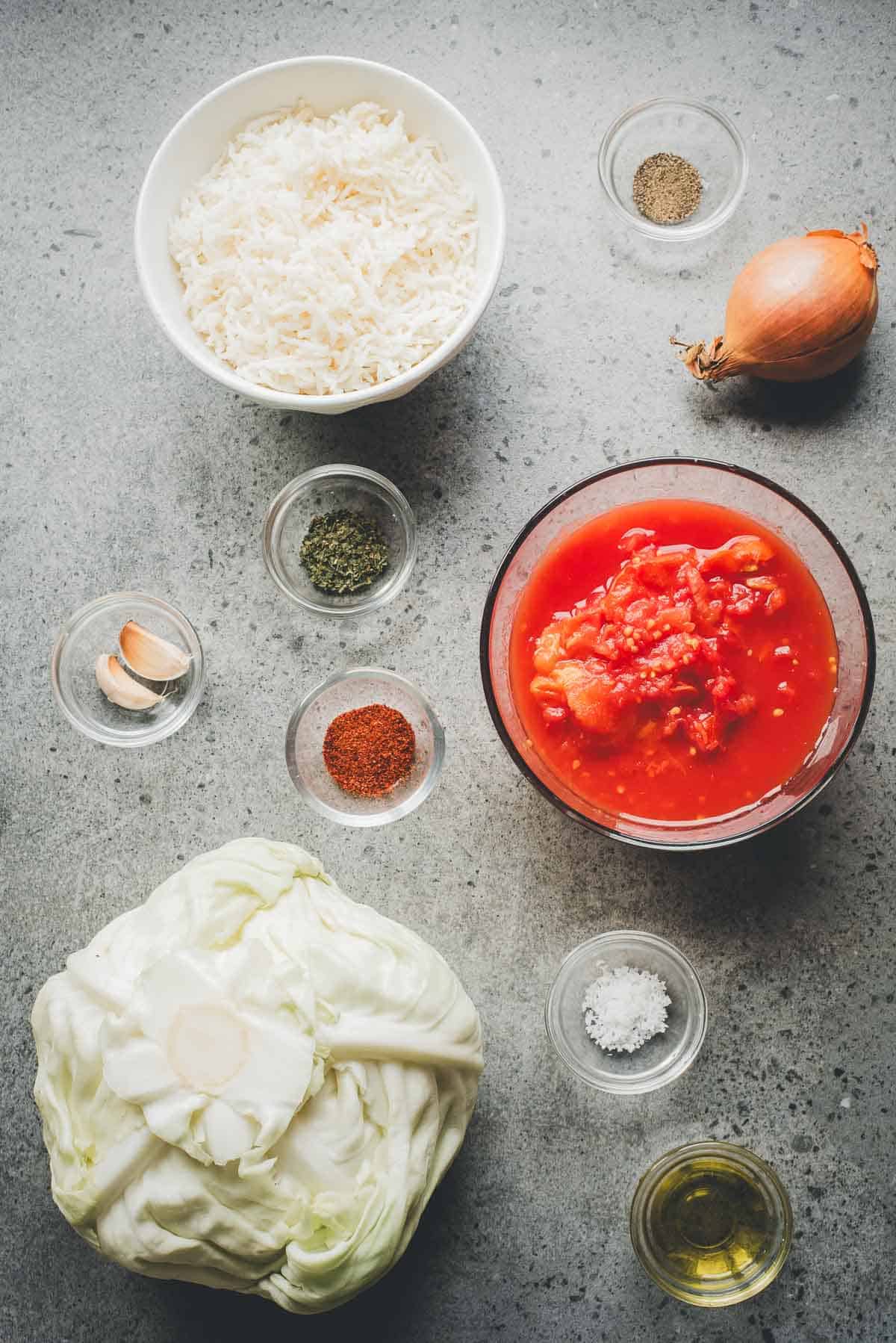 Overhead view of ingredients for Cabbage Rolls Casserole: rice, onion, canned tomatoes, cabbage, spices, garlic, and oil arranged on a gray surface.