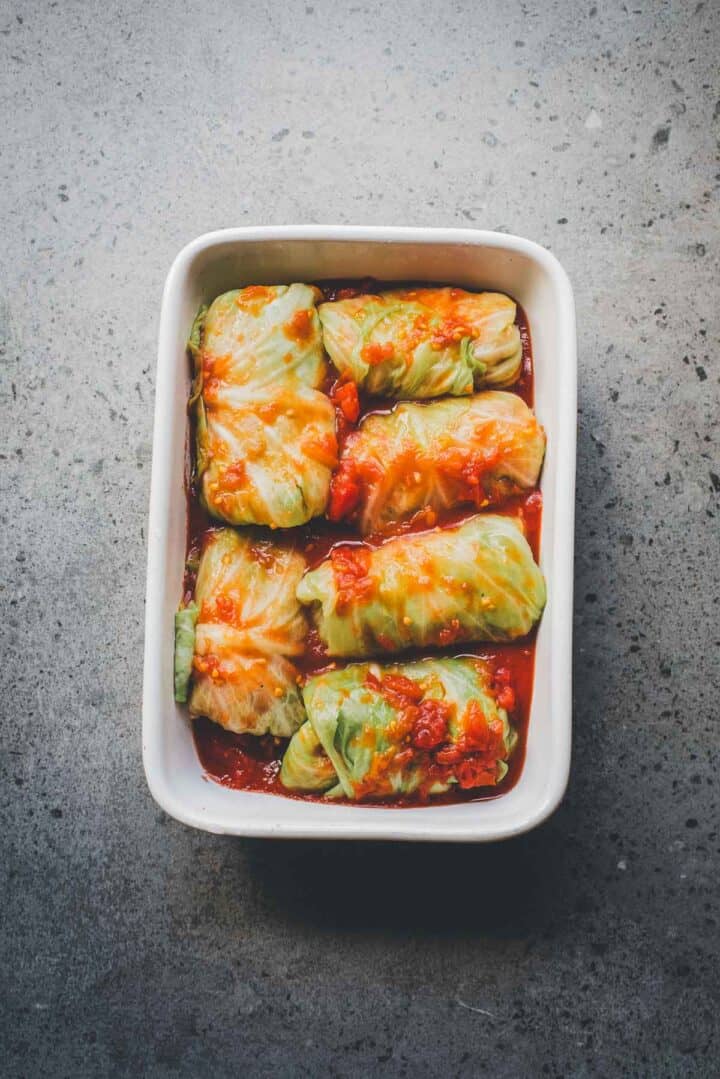 White dish with Cabbage Rolls Casserole, stuffed and baked in tomato sauce, arranged neatly on a gray speckled surface.