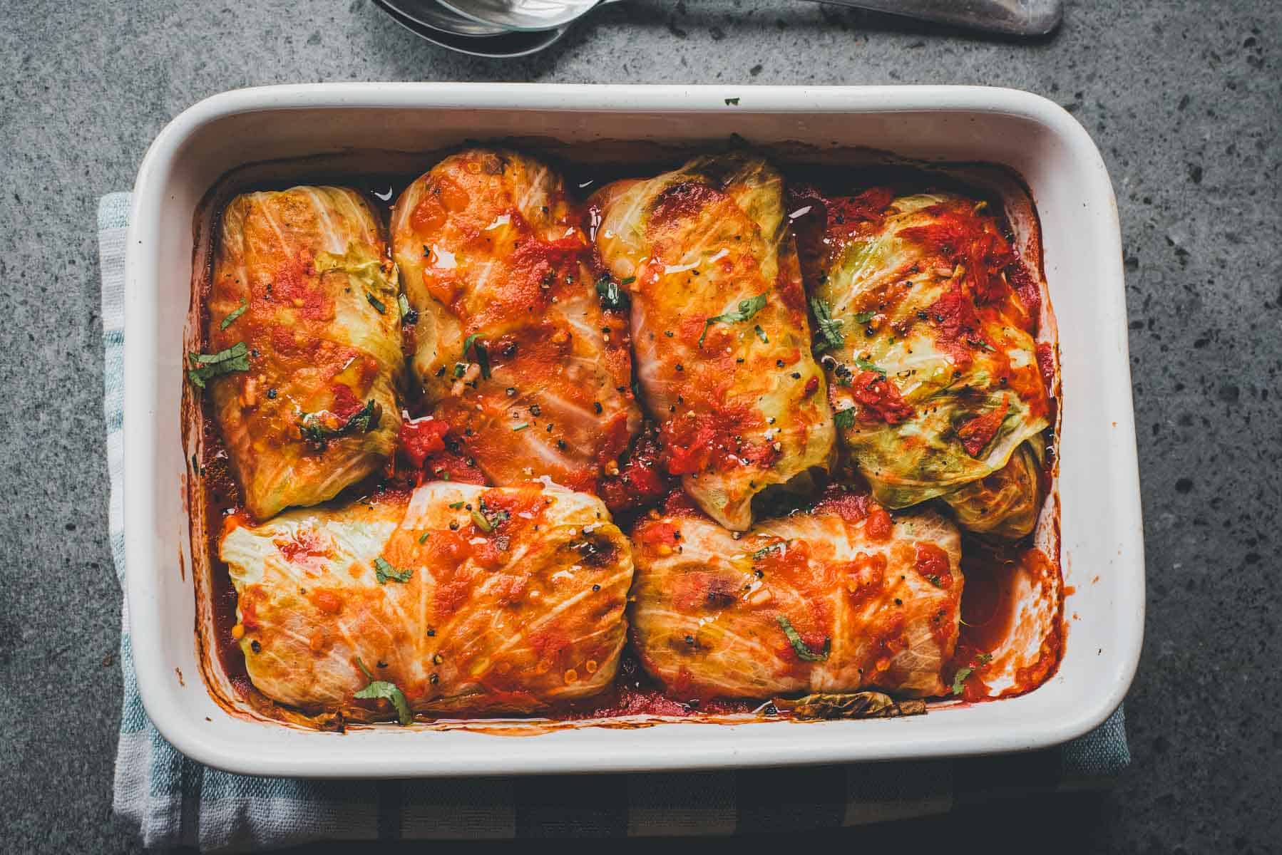 Baked Cabbage Rolls Casserole in tomato sauce is served in a white rectangular dish, garnished with fresh herbs.