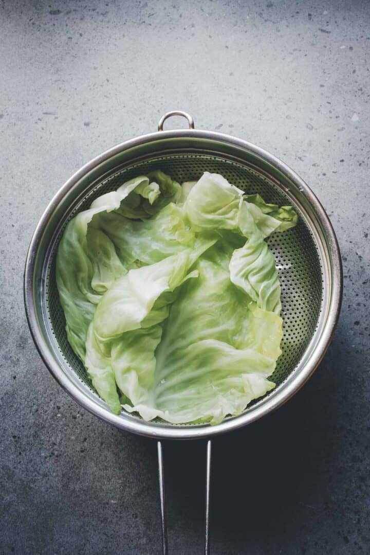 Large cabbage leaves in a metal strainer on a gray countertop, viewed from above&mdash;perfect for preparing your next Cabbage Rolls Casserole.