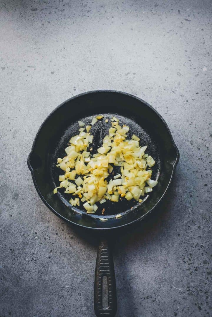 Chopped onions and garlic sizzle in a black skillet on a gray stone surface, the perfect base for a flavorful Cabbage Rolls Casserole.