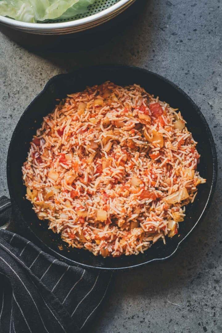 A skillet filled with cooked rice, tomatoes, vegetables, and savory flavors reminiscent of Cabbage Rolls Casserole sits on a gray counter beside a striped towel.