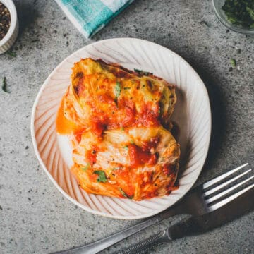 Two cabbage rolls topped with tomato sauce sit on a white plate, with a fork and knife beside the plate—perfect for anyone craving the comforting flavors of Cabbage Rolls Casserole.