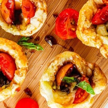 Four puff pastry tarts with cherry tomatoes, basil, and balsamic glaze make elegant appetizers on a wooden cutting board.