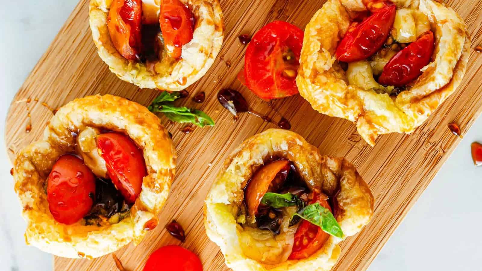 Four puff pastry tarts with cherry tomatoes, basil, and balsamic glaze make elegant appetizers on a wooden cutting board.