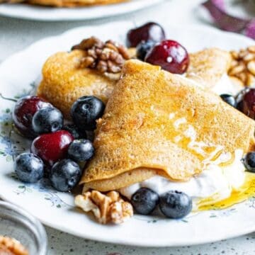 A plate with crepes, whipped cream, blueberries, cherries, walnuts, and honey on a white floral dish—perfect for adding to your collection of Easter brunch recipes.