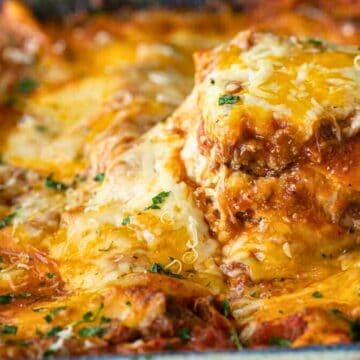 A slice of cheesy, baked lasagna being served from a pan, topped with herbs and melted cheese.