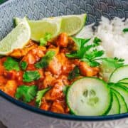 A bowl of Coconut Chicken Rice Bowls served with curry, rice, lime wedges, sliced cucumber, and a fresh cilantro garnish.