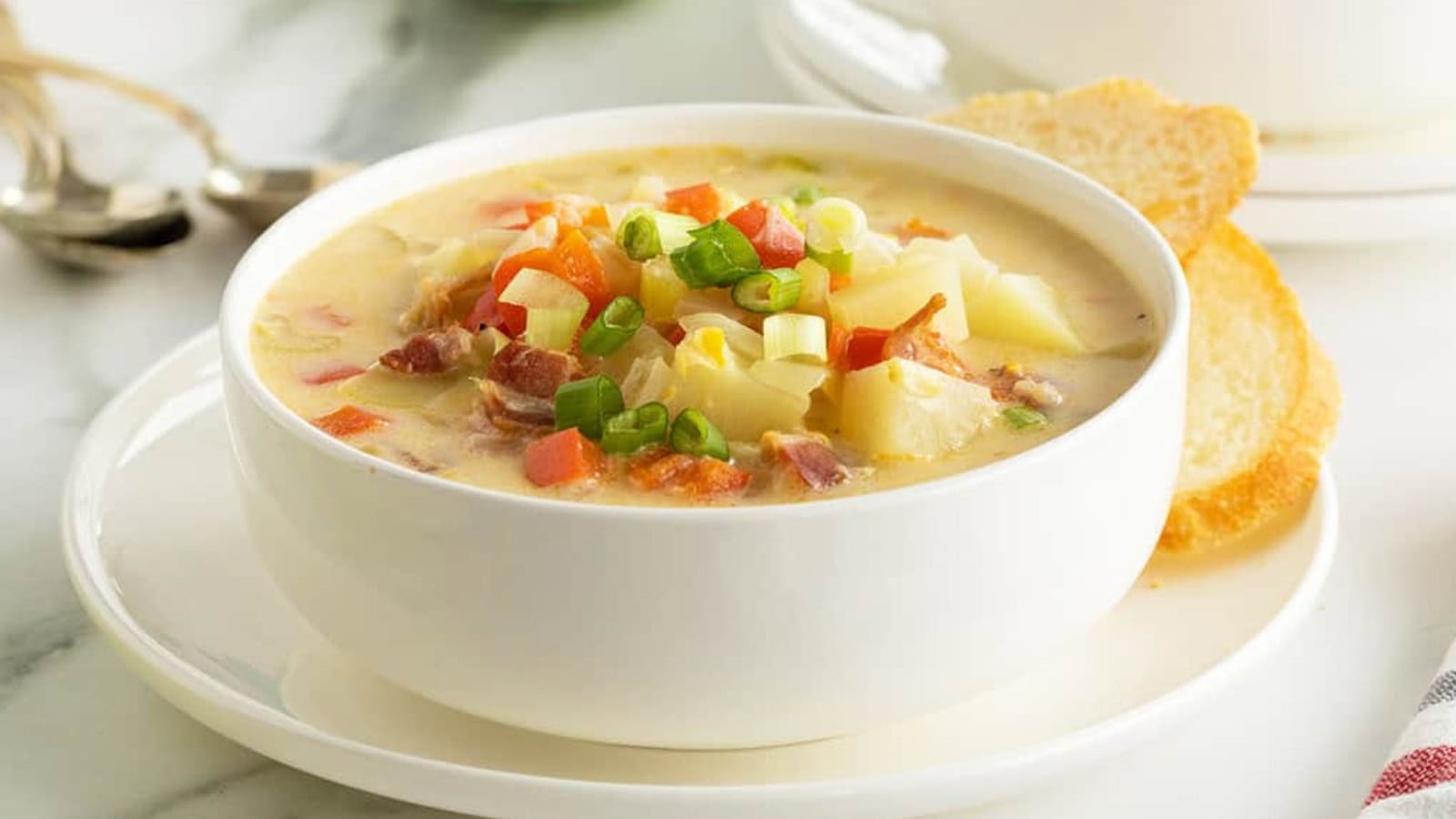 A bowl of creamy soup with vegetables and bacon, garnished with green onions, served with bread slices&mdash;a perfect choice for budget comfort recipes.