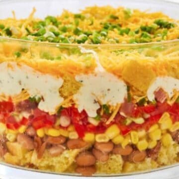 Layered salad in a glass bowl with cheese, green onions, chips, dressing, beans, corn, tomatoes, and lettuce.
