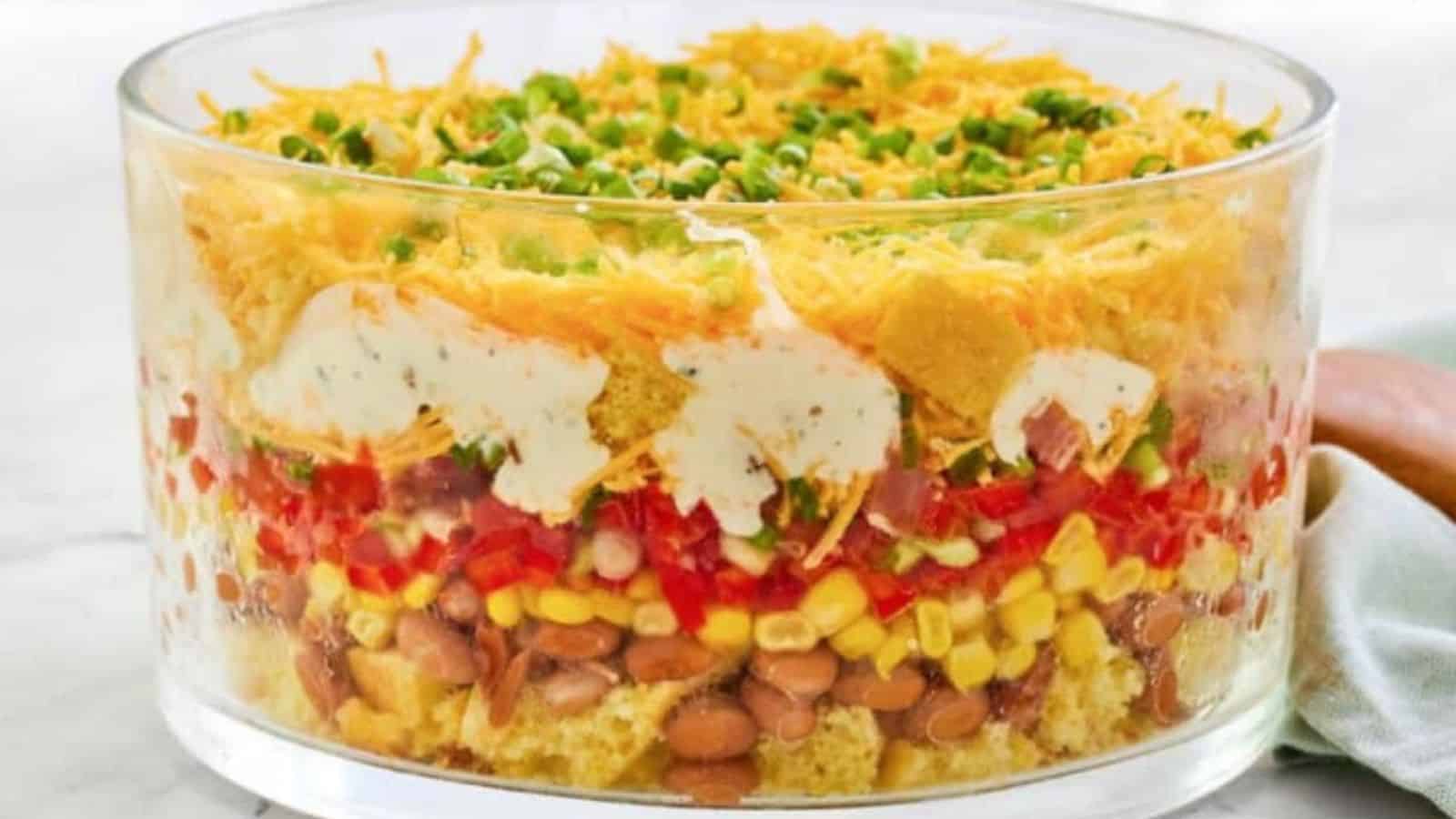 Layered salad in a glass bowl with cheese, green onions, chips, dressing, beans, corn, tomatoes, and lettuce.