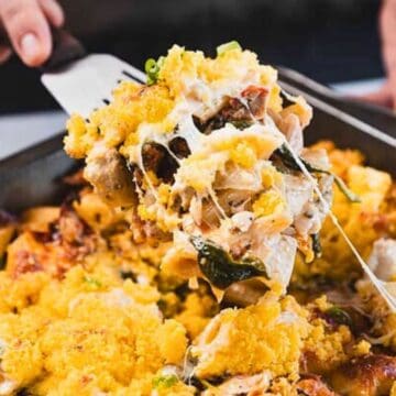 A hand serves a cheesy baked pasta casserole with a golden, crunchy topping from a pan.