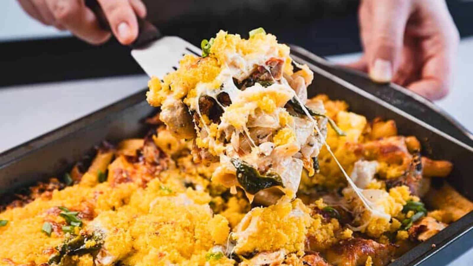 A close-up of a baked pasta dish in a rectangular metal pan with a golden breadcrumb topping, pieces of pasta, baked cheese, and green herbs visible.