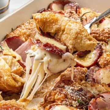 A close-up of a cheesy croissant bake with ham and apple slices being served from a baking dish.