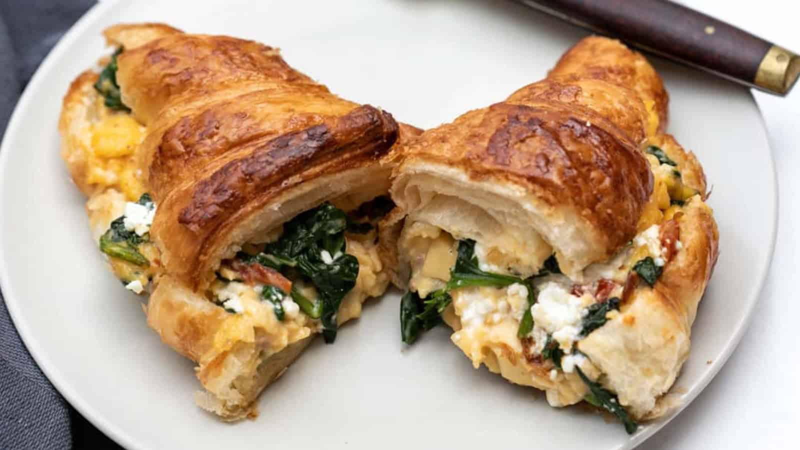 A croissant filled with scrambled eggs, spinach, and cheese sits on a white plate with a knife nearby—perfect for those who love easy brunch recipes.