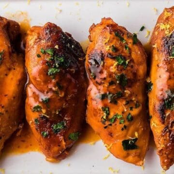 Four glazed chicken breasts garnished with parsley and lemon slices on a white plate.