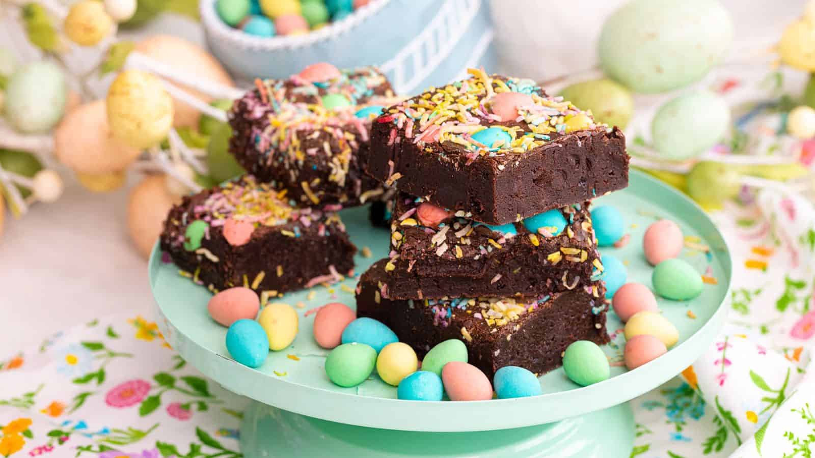 Chocolate brownies topped with colorful sprinkles and candies, stacked on a plate with candy eggs around them.