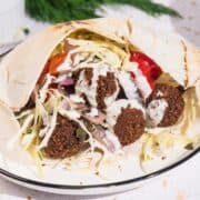 Falafel pita sandwich with Israeli Falafel Style Cabbage Salad, tomatoes, onions, and tahini sauce on a white plate.