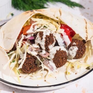 Falafel pita sandwich with Israeli Falafel Style Cabbage Salad, tomatoes, onions, and tahini sauce on a white plate.