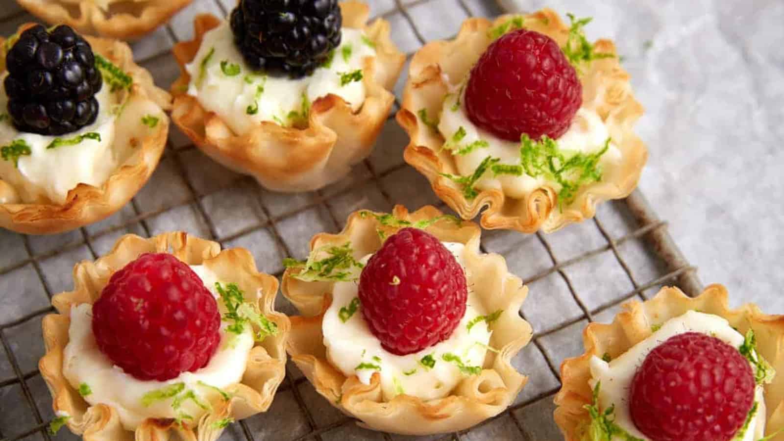 Mini tartlets make delightful appetizers, filled with creamy goodness and topped with raspberries, blackberries, and lime zest on a cooling rack.