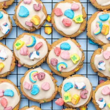 Cookies topped with white icing and colorful marshmallow cereal pieces on a cooling rack.