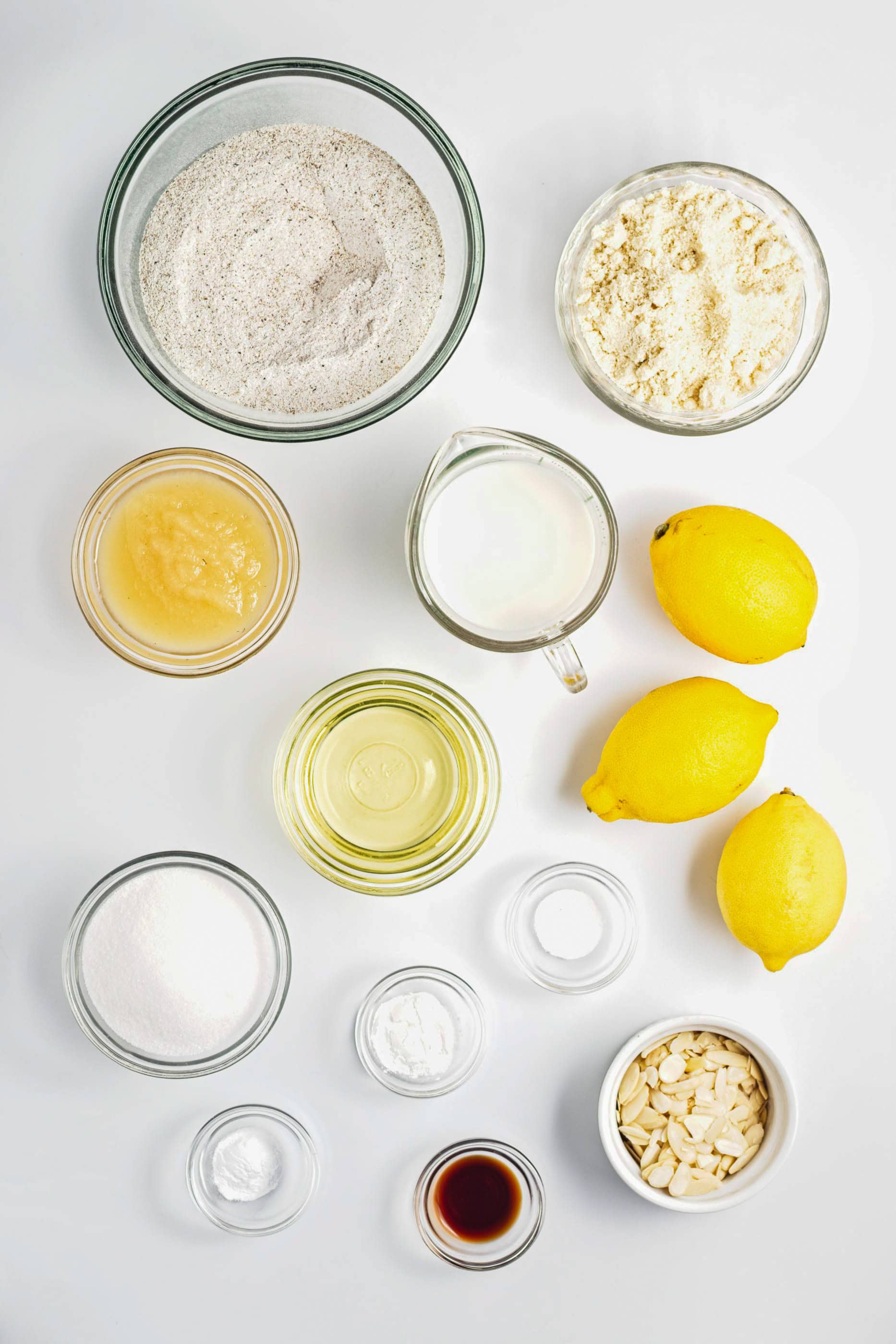 Baking ingredients arranged on a white surface, including lemons, flour, sugar, milk, oil, and small bowls of powders—perfect for creating a Gluten-Free Lemon Almond Loaf.