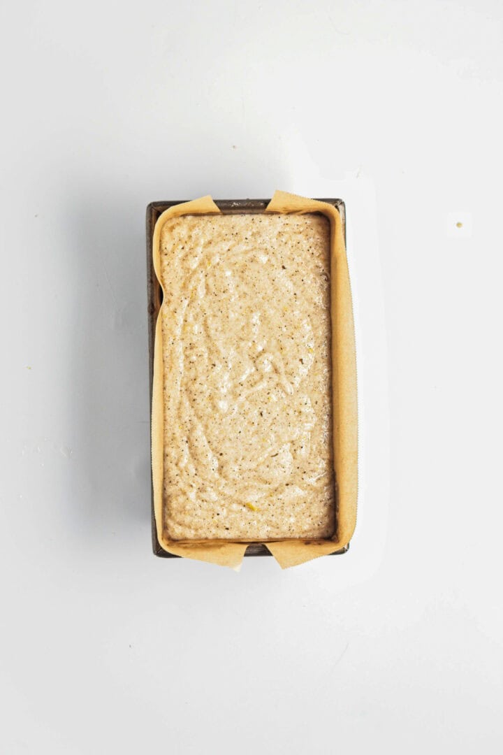 A loaf pan lined with parchment paper is filled with unbaked Gluten-Free Lemon Almond Loaf batter, ready to bake on a white surface.