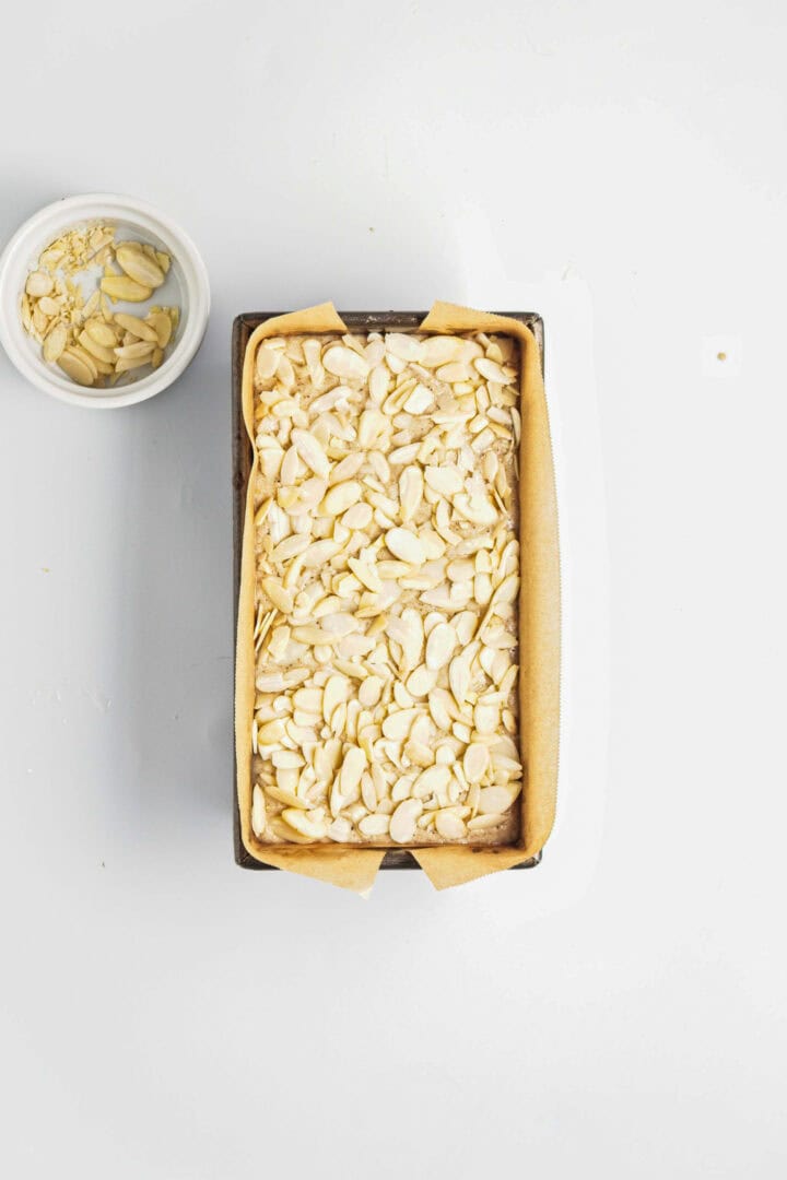 A Gluten-Free Lemon Almond Loaf in a loaf pan with sliced almonds on the batter, next to a small bowl of more sliced almonds on a white surface.