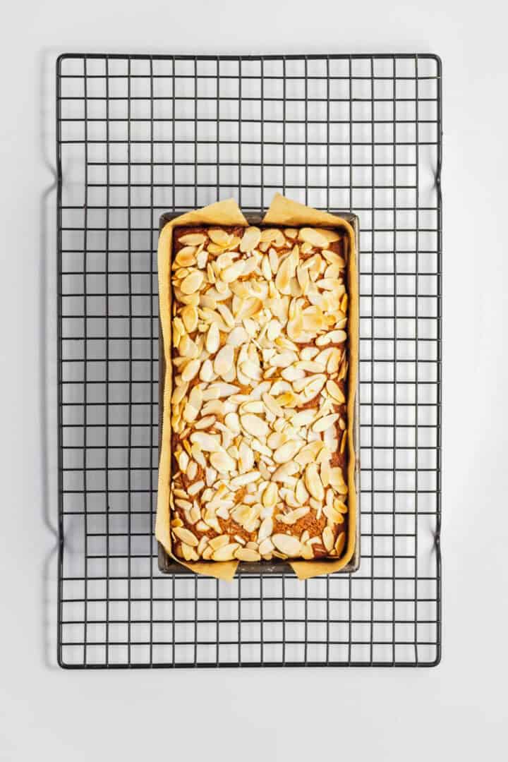 Gluten-Free Lemon Almond Loaf topped with sliced almonds in a parchment-lined pan on a black cooling rack, viewed from above.