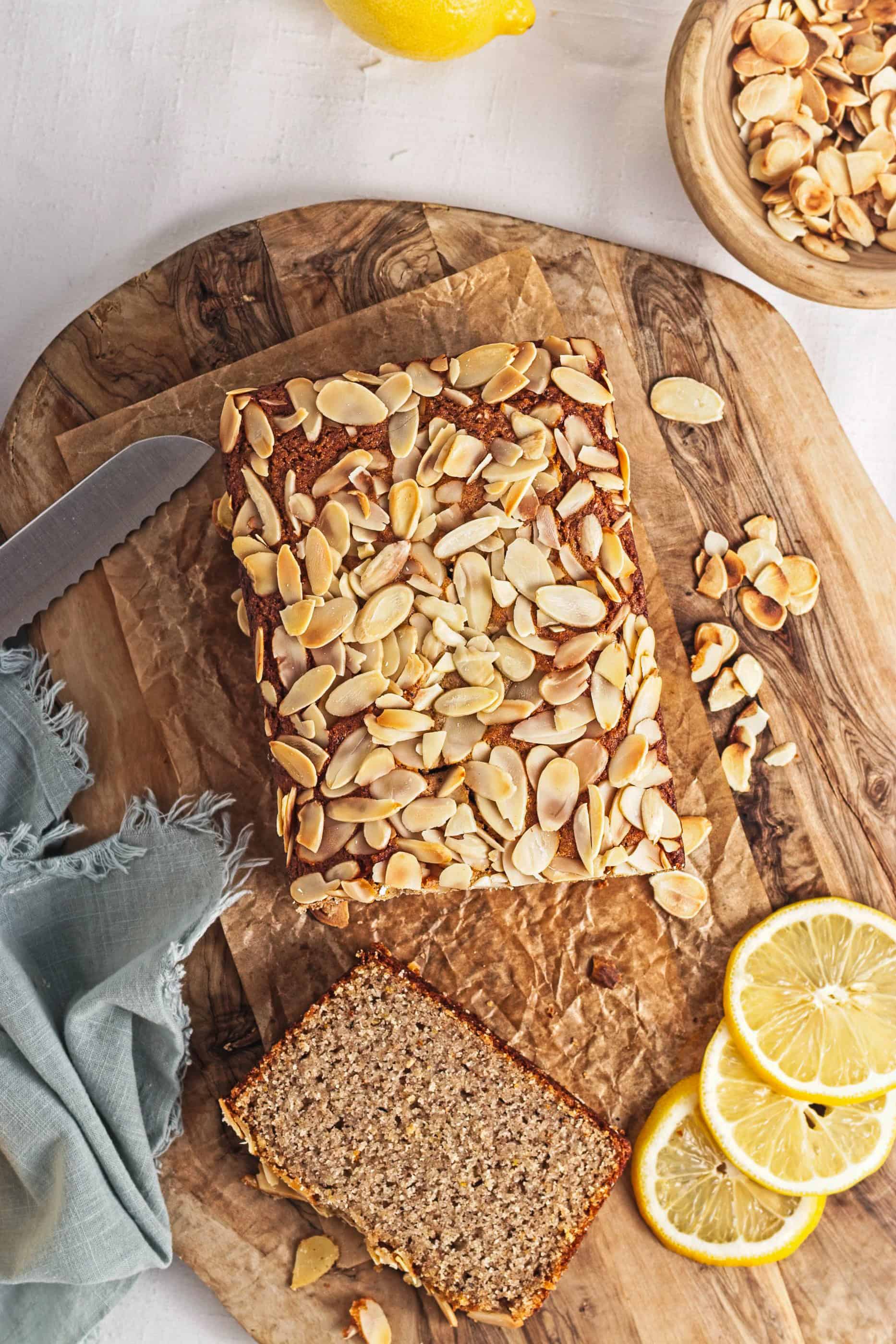 A Gluten-Free Lemon Almond Loaf topped with almonds sits on a wooden board, with a slice cut, lemon slices, and a bowl of almonds nearby.
