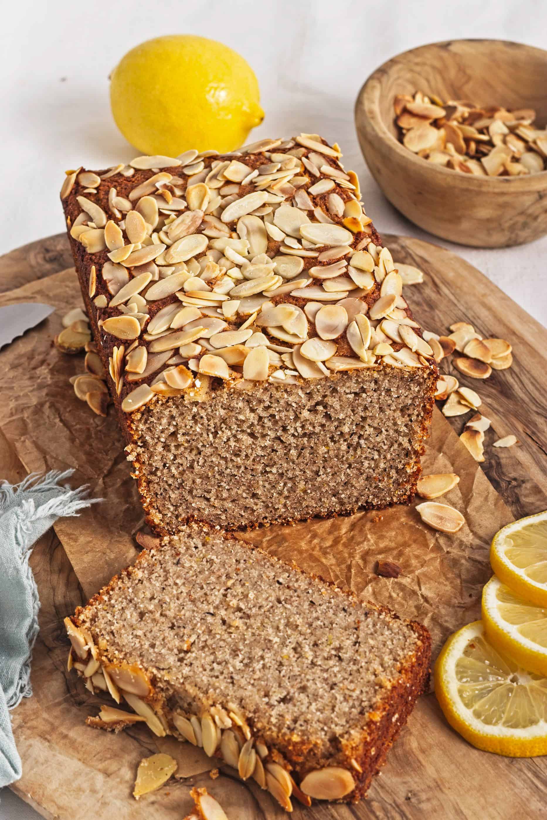 A Gluten-Free Lemon Almond Loaf with sliced almonds on top, one slice cut, and fresh lemon slices nearby.