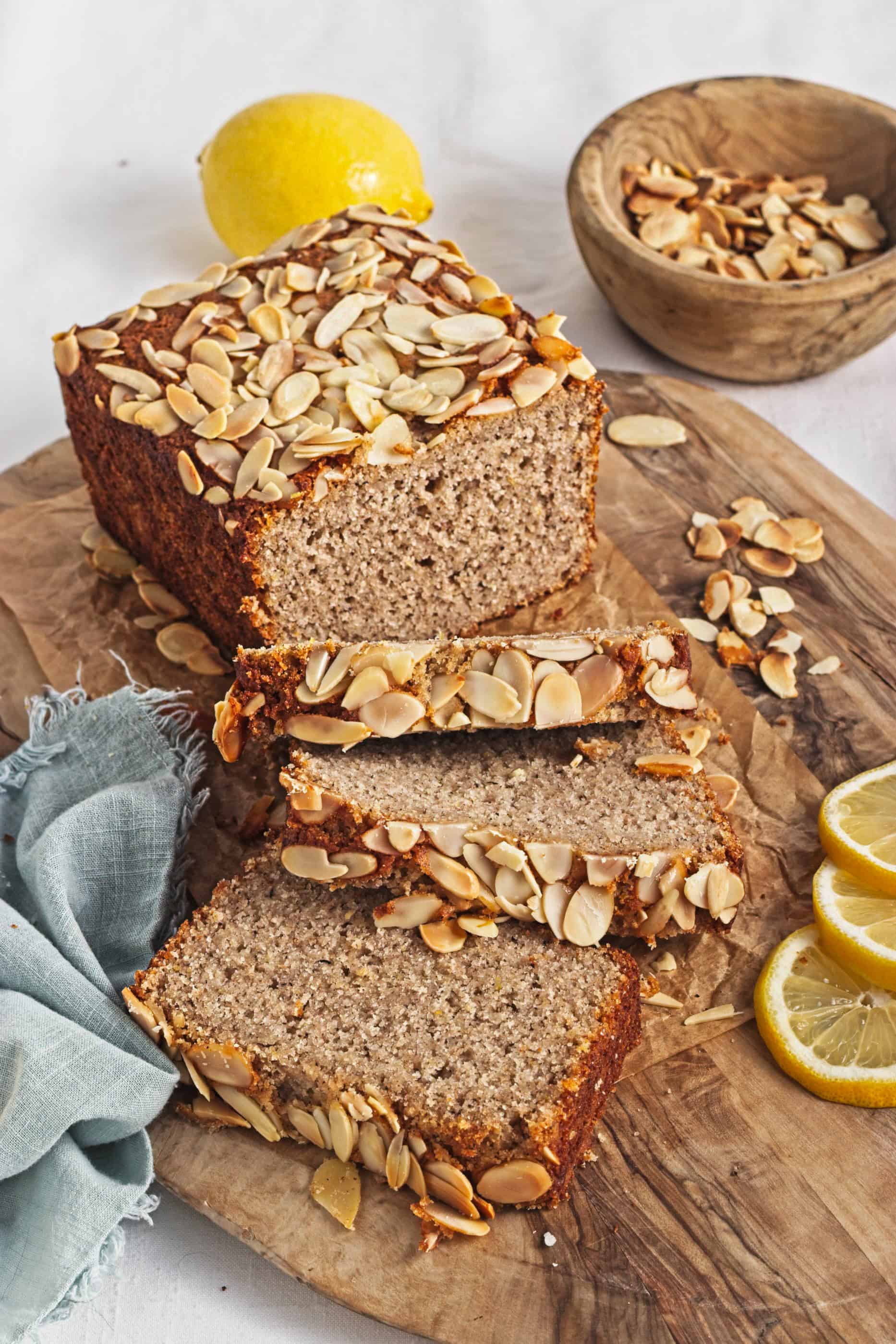 A Gluten-Free Lemon Almond Loaf, topped with almond flakes and lemon, is sliced and served on a wooden board.