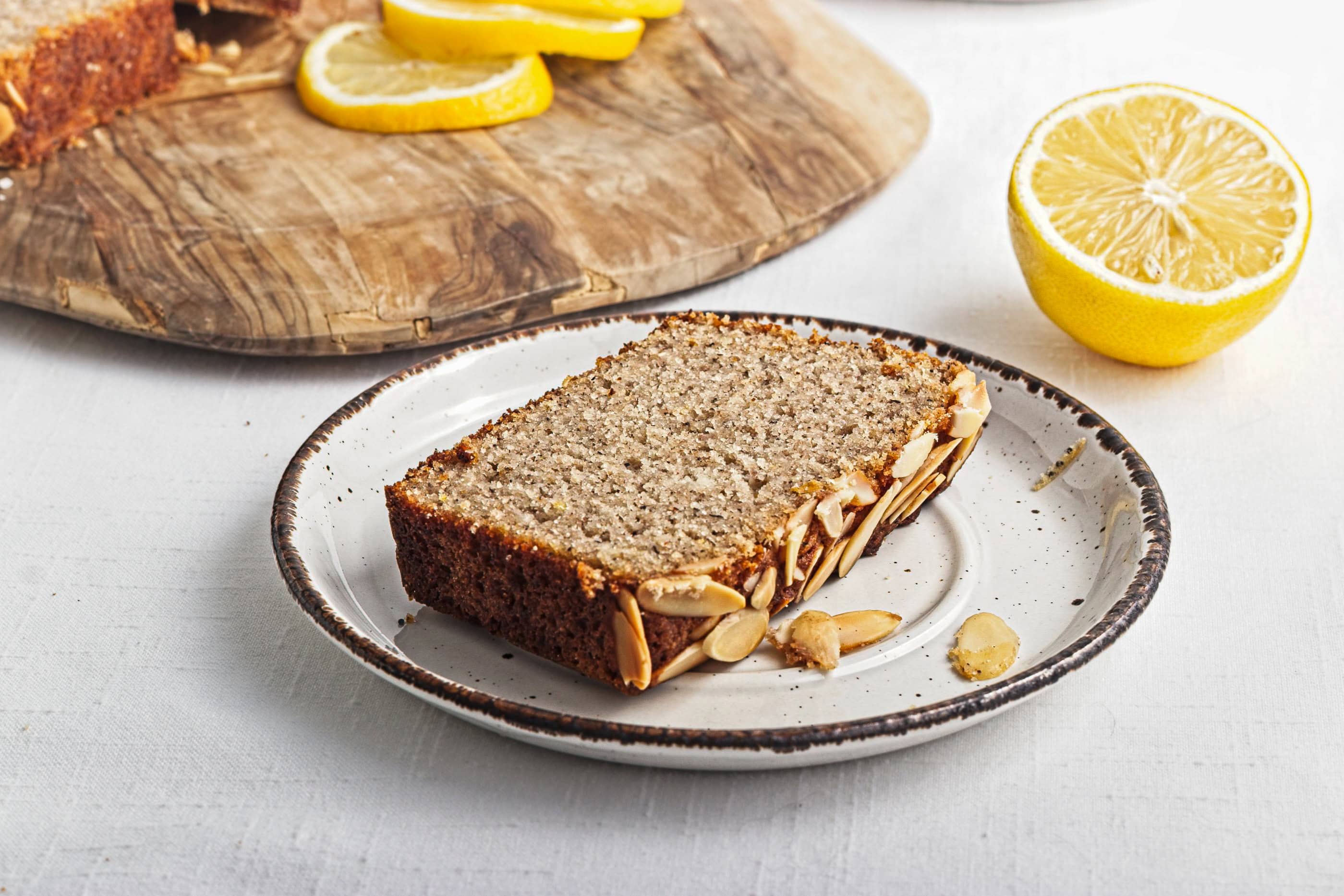 A slice of Gluten-Free Lemon Almond Loaf on a plate, topped with nuts, with lemon slices and a halved lemon nearby.