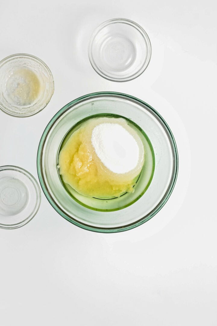 A glass bowl with applesauce and sugar, surrounded by three empty glass bowls on a white background, perfect for preparing a Gluten-Free Lemon Almond Loaf.