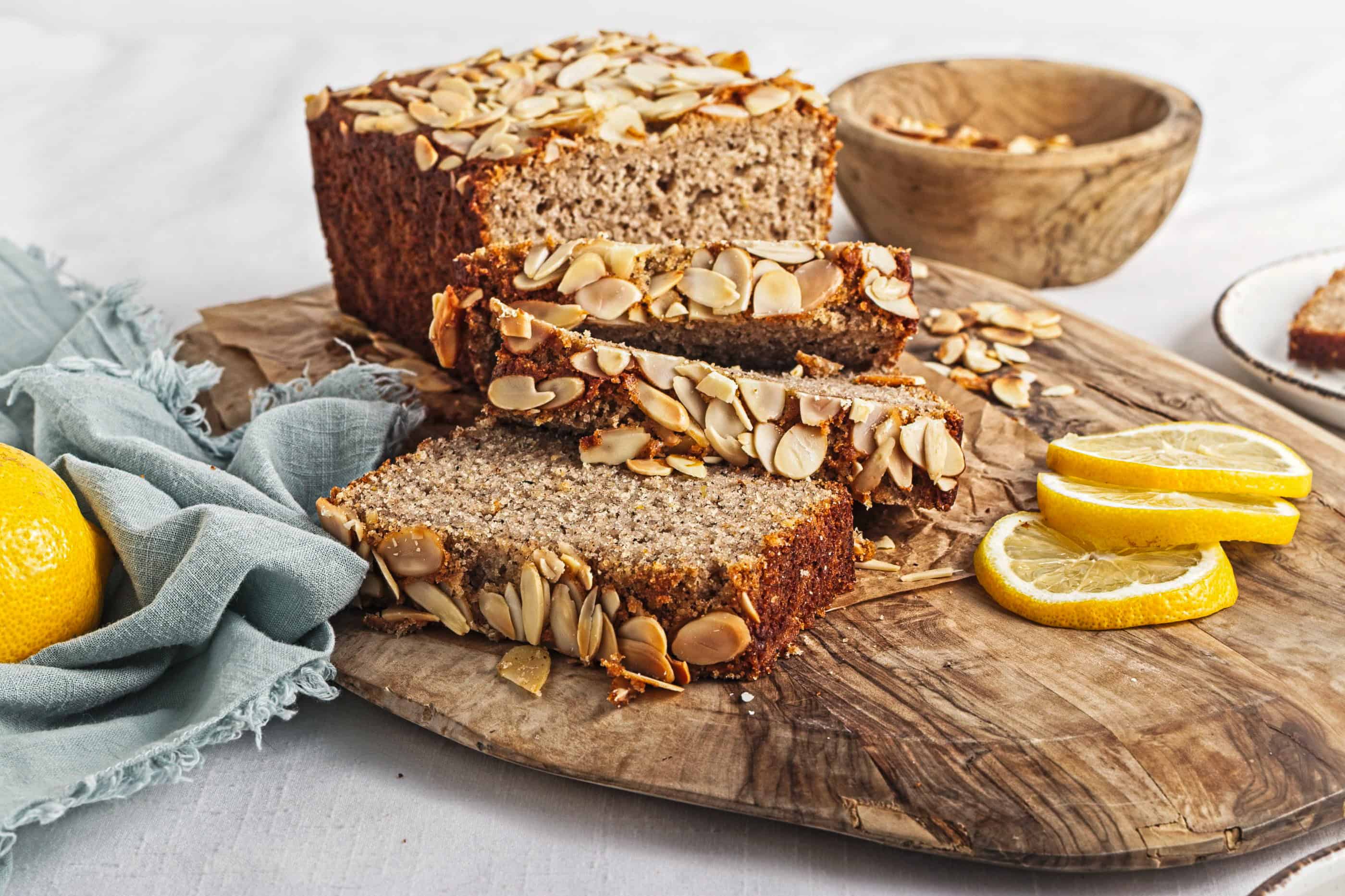Gluten-Free Lemon Almond Loaf topped with sliced almonds, served on a wooden board alongside lemon slices and a blue cloth nearby.