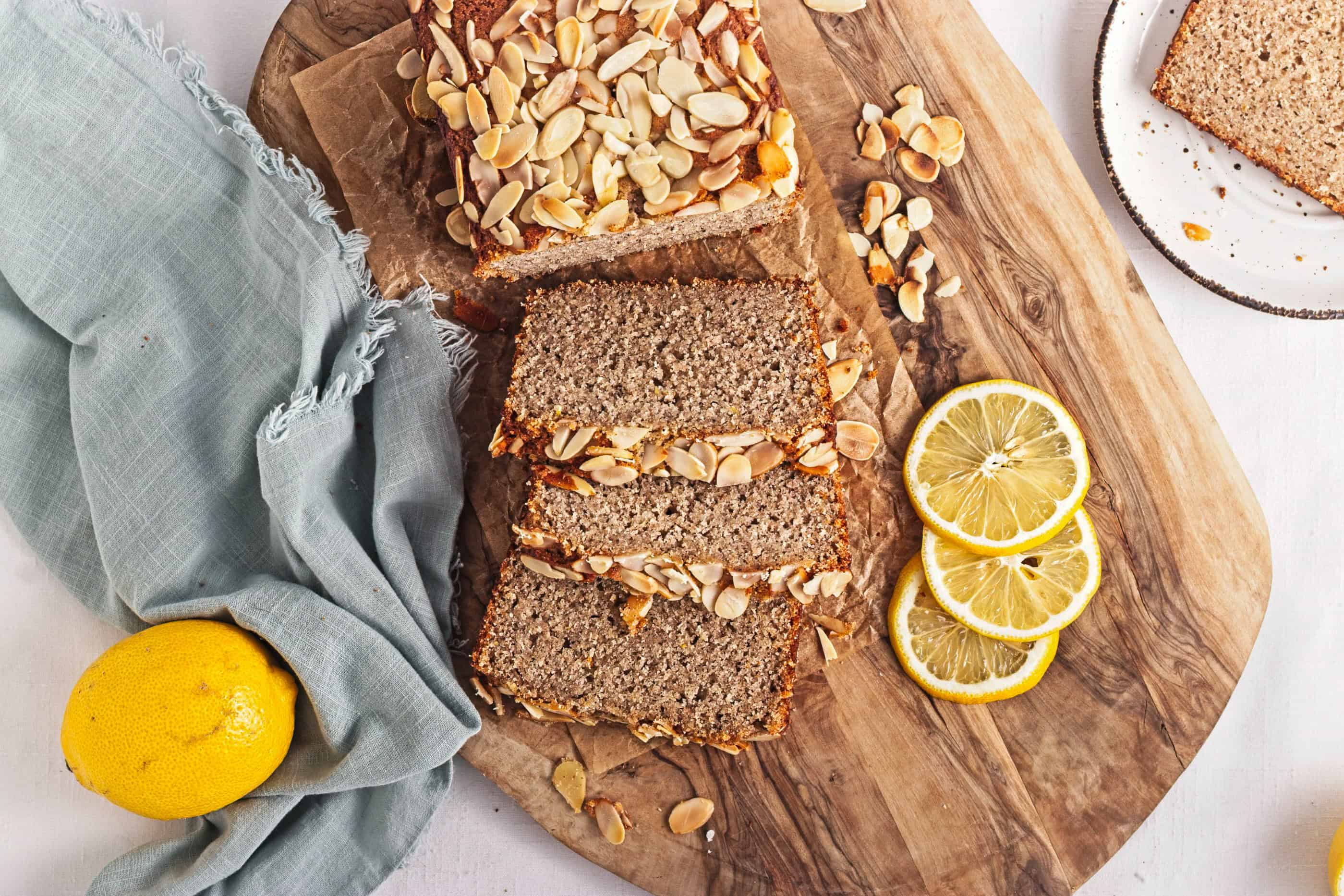 Gluten-Free Lemon Almond Loaf sliced on a wooden board with lemon slices, a whole lemon, and a blue napkin.