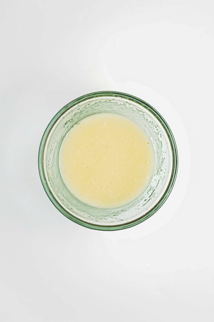 A glass bowl filled with a creamy, light yellow liquid on a white background, perfect for drizzling over a Gluten-Free Lemon Almond Loaf.