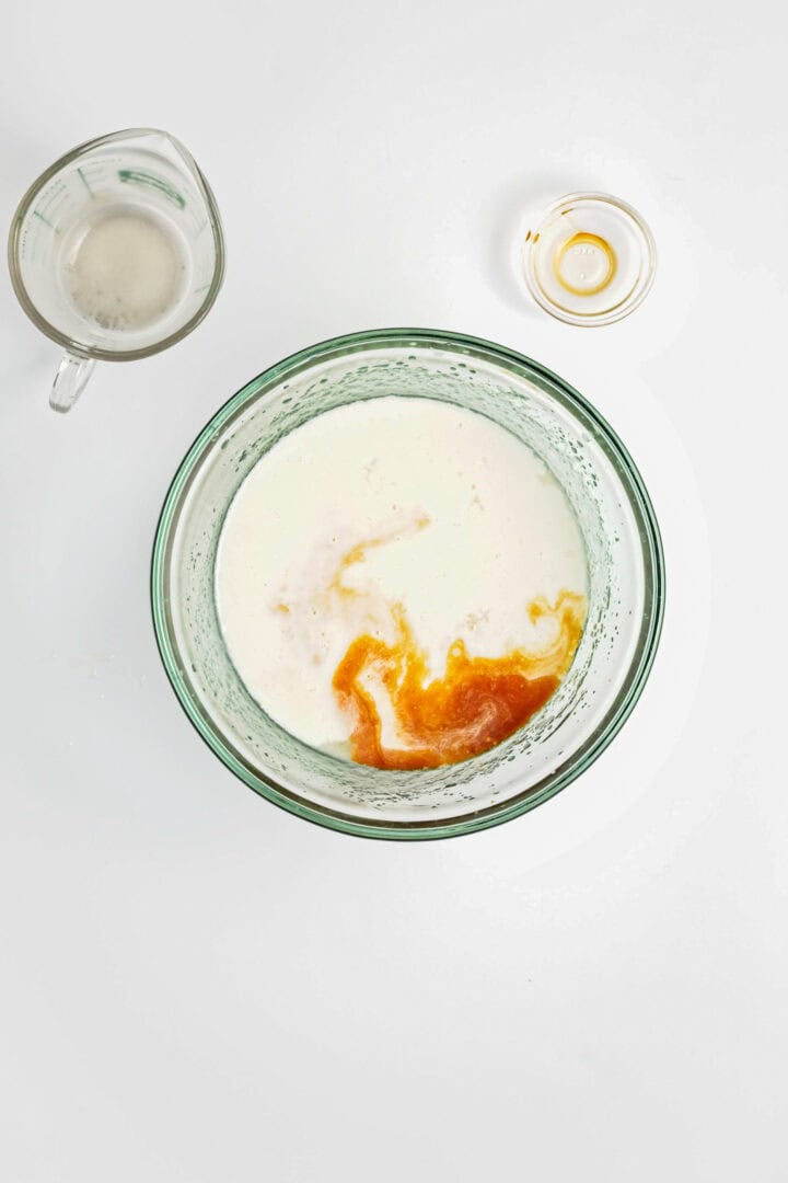 A glass bowl with milk and vanilla, ready to be mixed for a Gluten-Free Lemon Almond Loaf, sits beside an empty measuring cup and a small dish on a white surface.
