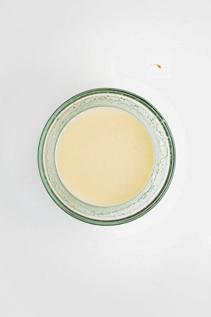 A glass bowl filled with a creamy, light beige liquid—perfect for drizzling over a Gluten-Free Lemon Almond Loaf—rests on a white background.