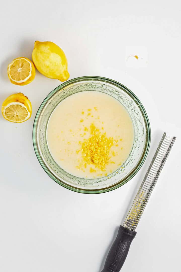 A bowl of batter with lemon zest on top, next to lemons and a zester on a white surface—perfect for making a Gluten-Free Lemon Almond Loaf.
