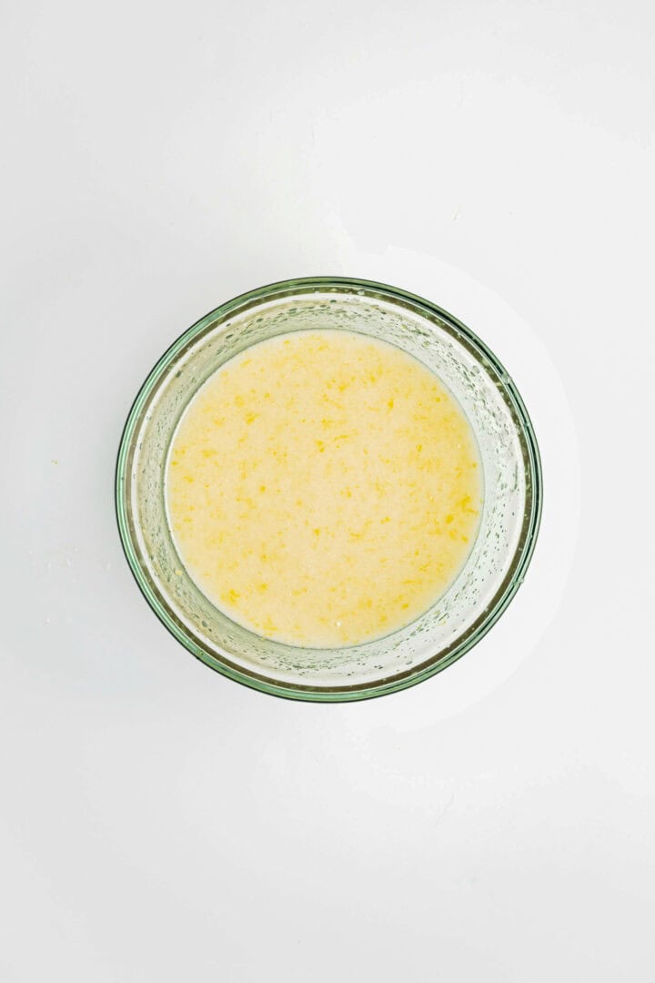 A glass bowl filled with a pale yellow liquid mixture, ready to become a delicious Gluten-Free Lemon Almond Loaf, viewed from above on a white background.