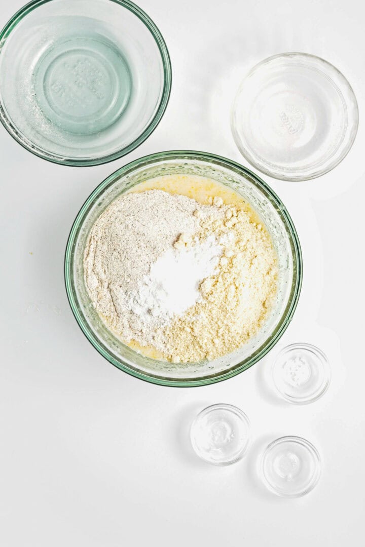 Bowls with flour, baking soda, and other baking ingredients for a Gluten-Free Lemon Almond Loaf on a white surface, viewed from above.