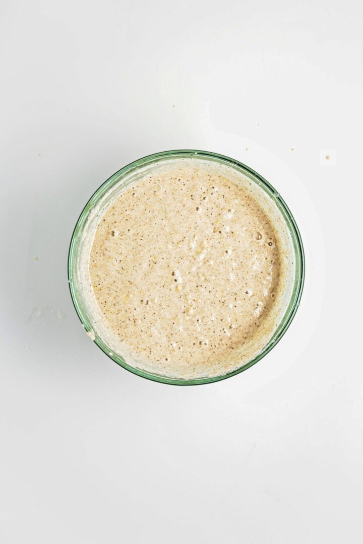 A glass bowl filled with bubbly sourdough starter, perfect for baking treats like a Gluten-Free Lemon Almond Loaf, sits on a white background, viewed from above.