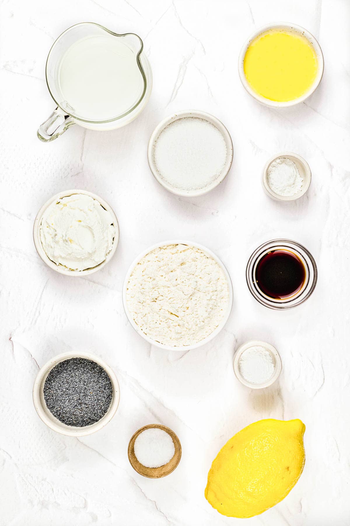 Overhead view of baking ingredients including milk, butter, flour, sugar, poppy seeds, lemon, and vanilla on white background.