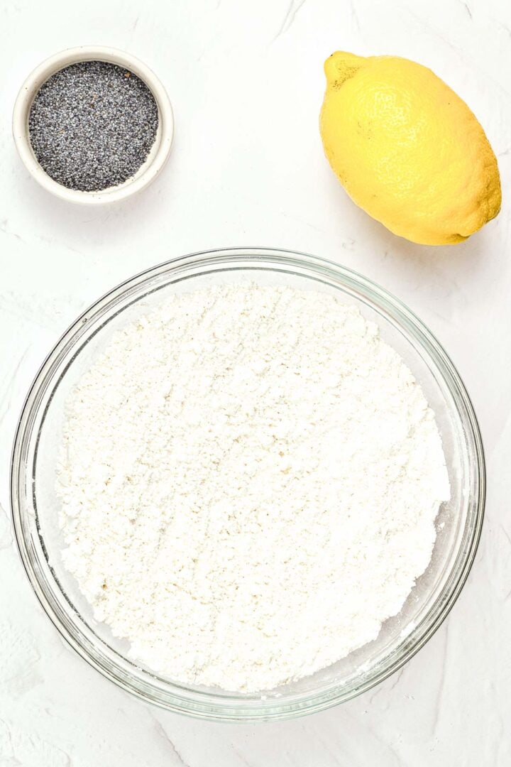 A bowl of flour, a lemon, and a small dish of poppy seeds on a white surface.