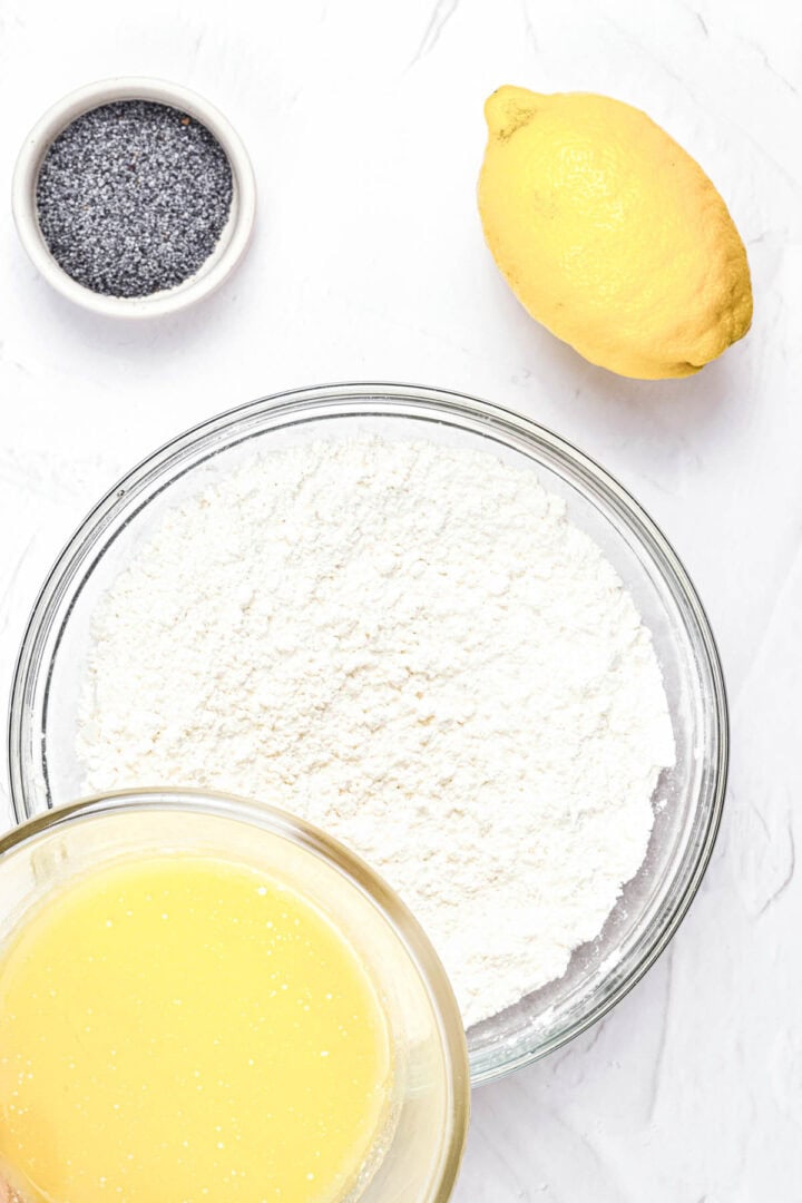 A lemon, poppy seeds, flour, and melted butter in bowls on a white surface.